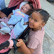 Yasmine participe au concours pour gagner de l'argent avec cette photo : toddler, child, stroller, blanket, plush_toy, smiling, happy, outdoor, concrete, car_tire, person, clothing, shorts, earrings, curly_hair, hand, footwear, sibling, family, cute