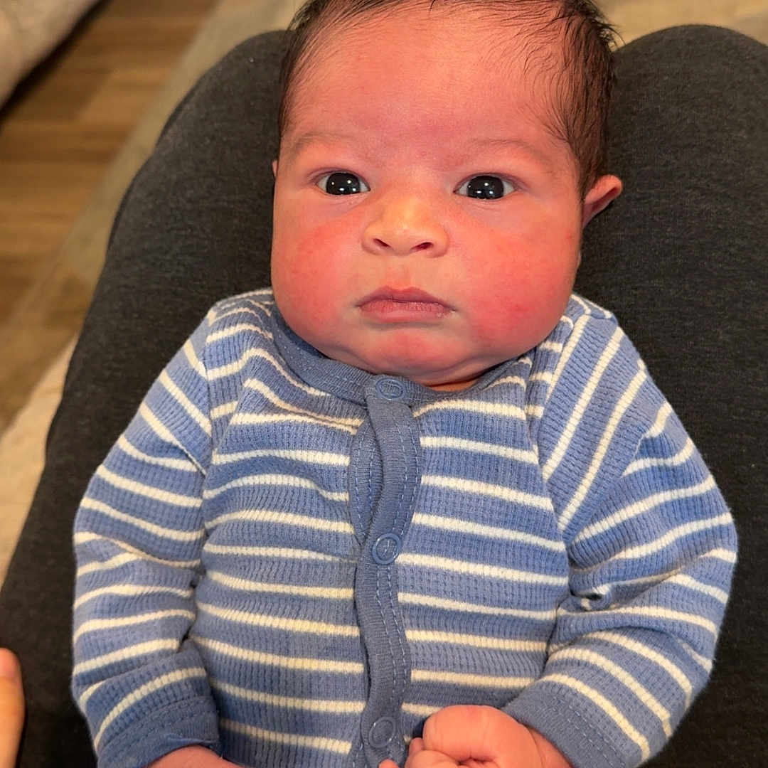 Karter joined the competition — help win amazing prizes! baby, carpet, child, cute, expression, face, fist, floor, furniture, hand, home, indoor, infant, looking_at_camera, newborn, onesie, person, portrait, sitting, striped_clothing