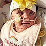 baby, infant, headband, flower, bib, nasal_tube, medical_device, close_up, face, expression, lying_down, pillow, pattern, butterfly, yellow, pink, clothing, person, newborn, soft_light
