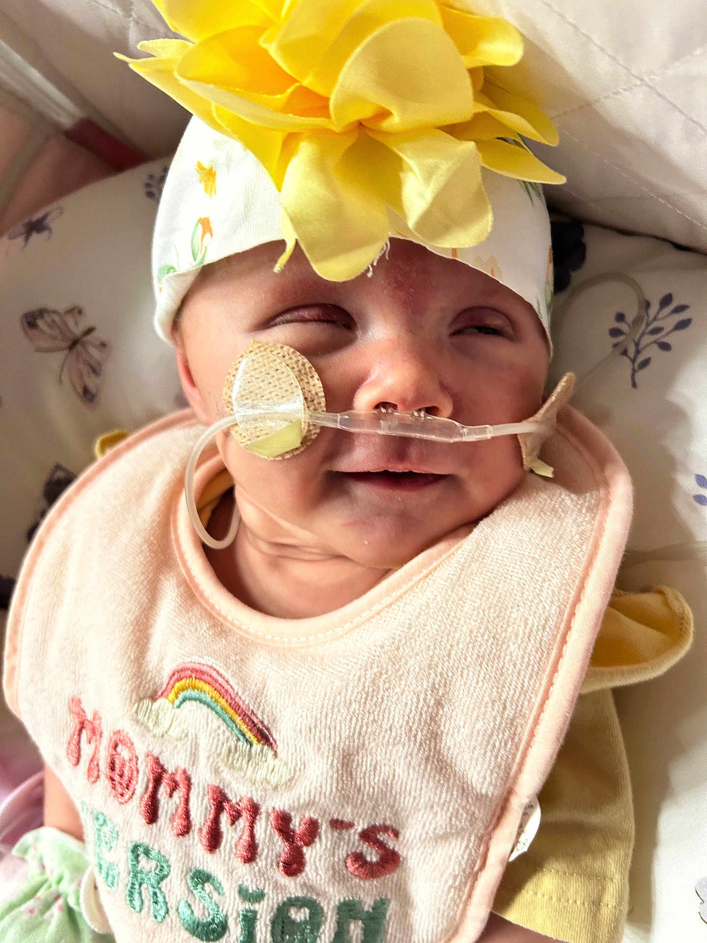 Calianna joined the competition — help win amazing prizes! baby, infant, headband, flower, bib, nasal_tube, medical_device, close_up, face, expression, lying_down, pillow, pattern, butterfly, yellow, pink, clothing, person, newborn, soft_light