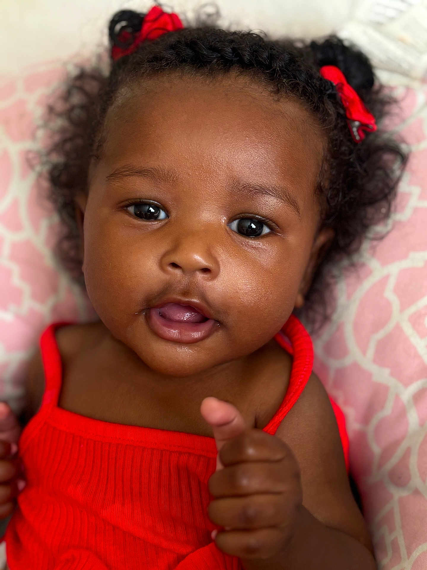 Areya joined the competition — help win amazing prizes! baby, child, curly_hair, red_bow, red_clothing, pointing_finger, close_up, portrait, cute, infant, pink_background, smiling, happy, face, skin, person, young, indoors, expression, headshot