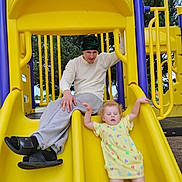 Sophia joined the competition — help win amazing prizes! activity, beanie, casual_clothing, child, colorful_hearts, falling, feet, fun, handrails, man, outdoor, park, person, play, playground, recreation, slide, trees, yellow_dress, yellow_slide