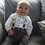 baby, smiling, sitting, couch, shirt, suspenders, pants, socks, dinosaur_print, window, pillows, happy, indoors, child, face, person, furniture, cute, young, comfortable