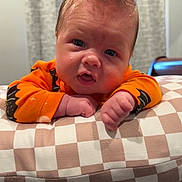 Lincoln is registered to the contest to win money with this photo: baby, infant, child, face, hair, mohawk, orange_clothing, bat_pattern, cushion, checkered_pattern, indoor, curtains, table, skin, expression, portrait, closeup, hands, furniture, floor