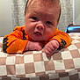 baby, infant, child, face, hair, mohawk, orange_clothing, bat_pattern, cushion, checkered_pattern, indoor, curtains, table, skin, expression, portrait, closeup, hands, furniture, floor