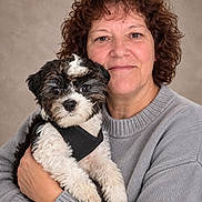 Zippy is registered to the contest to win money with this photo: adorable, black_and_white, companion, cuddling, curly_hair, dog, face, fluffy, harness, holding, indoors, person, pet, portrait, puppy, selfie, smiling, studio, sweater, woman