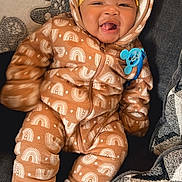 Antonio is registered to the contest to win money with this photo: baby, smiling, onesie, hood, ears, brown, white_patterns, sitting, couch, pillows, cozy, happy, child, indoor, cute, clothing, warm, headwear, sock, pacifier_clip