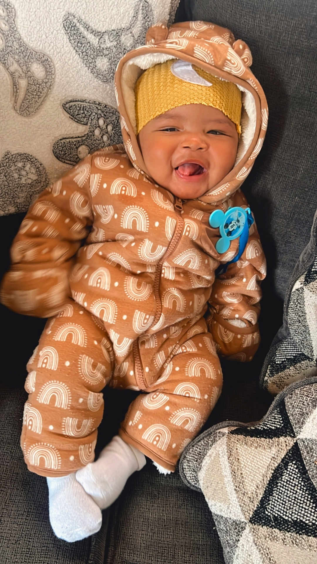Antonio is registered to the contest to win money with this photo: baby, smiling, onesie, hood, ears, brown, white_patterns, sitting, couch, pillows, cozy, happy, child, indoor, cute, clothing, warm, headwear, sock, pacifier_clip