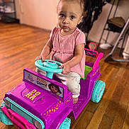 Melina is registered to the contest to win money with this photo: child, toddler, toy_car, indoor, wooden_floor, pink_clothing, purple_toy, teal_wheels, steering_wheel, serious_expression, home_interior, sitting, floor, person, play, cute, young_child, vehicle, flooring, furniture