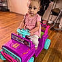 child, toddler, toy_car, indoor, wooden_floor, pink_clothing, purple_toy, teal_wheels, steering_wheel, serious_expression, home_interior, sitting, floor, person, play, cute, young_child, vehicle, flooring, furniture