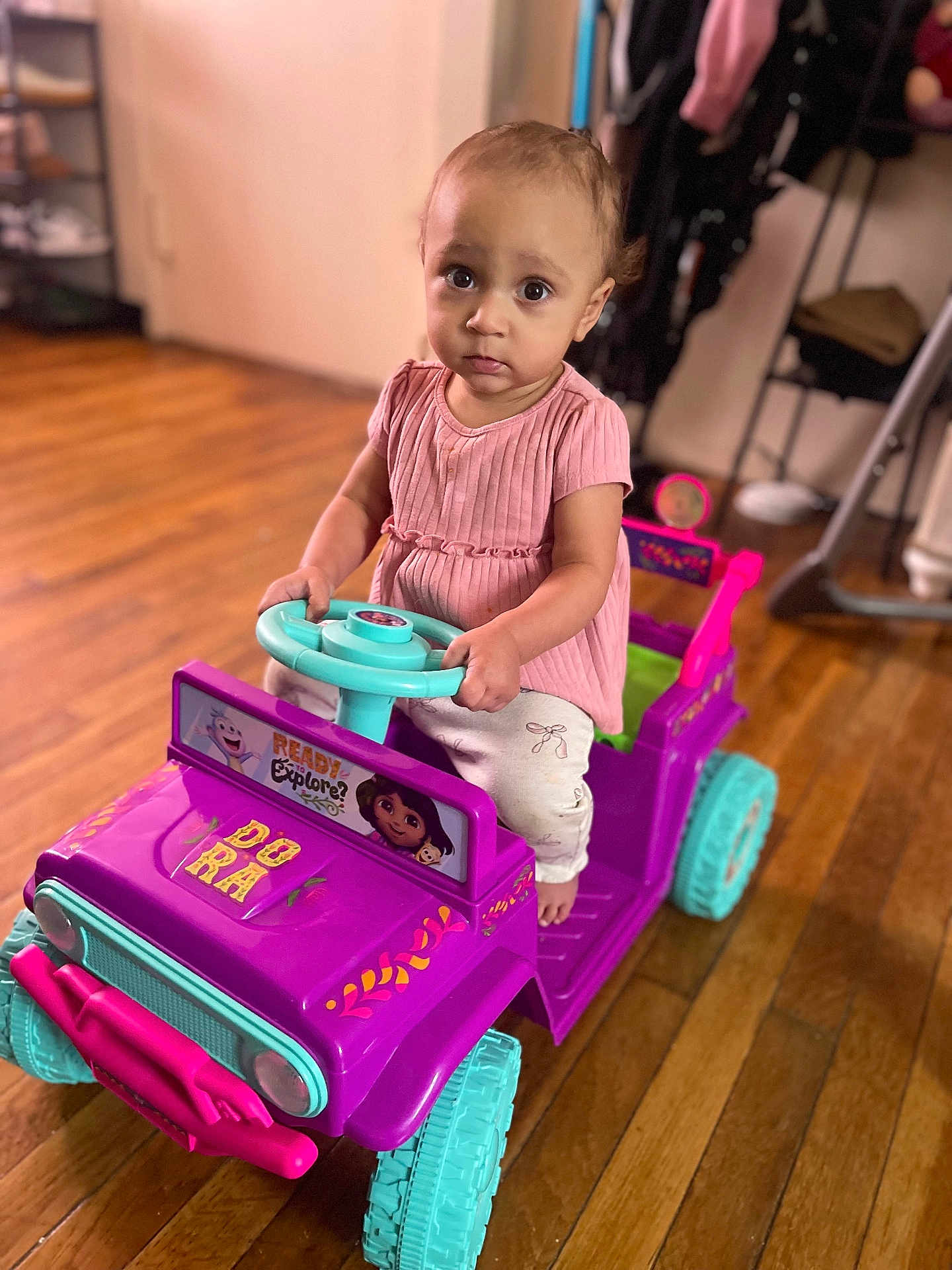 Melina is registered to the contest to win money with this photo: child, toddler, toy_car, indoor, wooden_floor, pink_clothing, purple_toy, teal_wheels, steering_wheel, serious_expression, home_interior, sitting, floor, person, play, cute, young_child, vehicle, flooring, furniture
