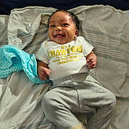 Xavier joined the competition — help win amazing prizes! baby, bear_slippers, bed, blanket, blanket_blue, child, clothing, cute, face, footwear, hands, happy, indoors, infant, lying_down, pants, person, shirt, smiling, text_on_shirt