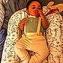 baby, infant, pacifier, onesie, suspenders, baby_lounger, dinosaur_print, pillow, blanket, bed, socks, hand, curly_hair, cheeks, indoor, soft_lighting, neutral_expression, cozy, red_pacifier_clip, portrait