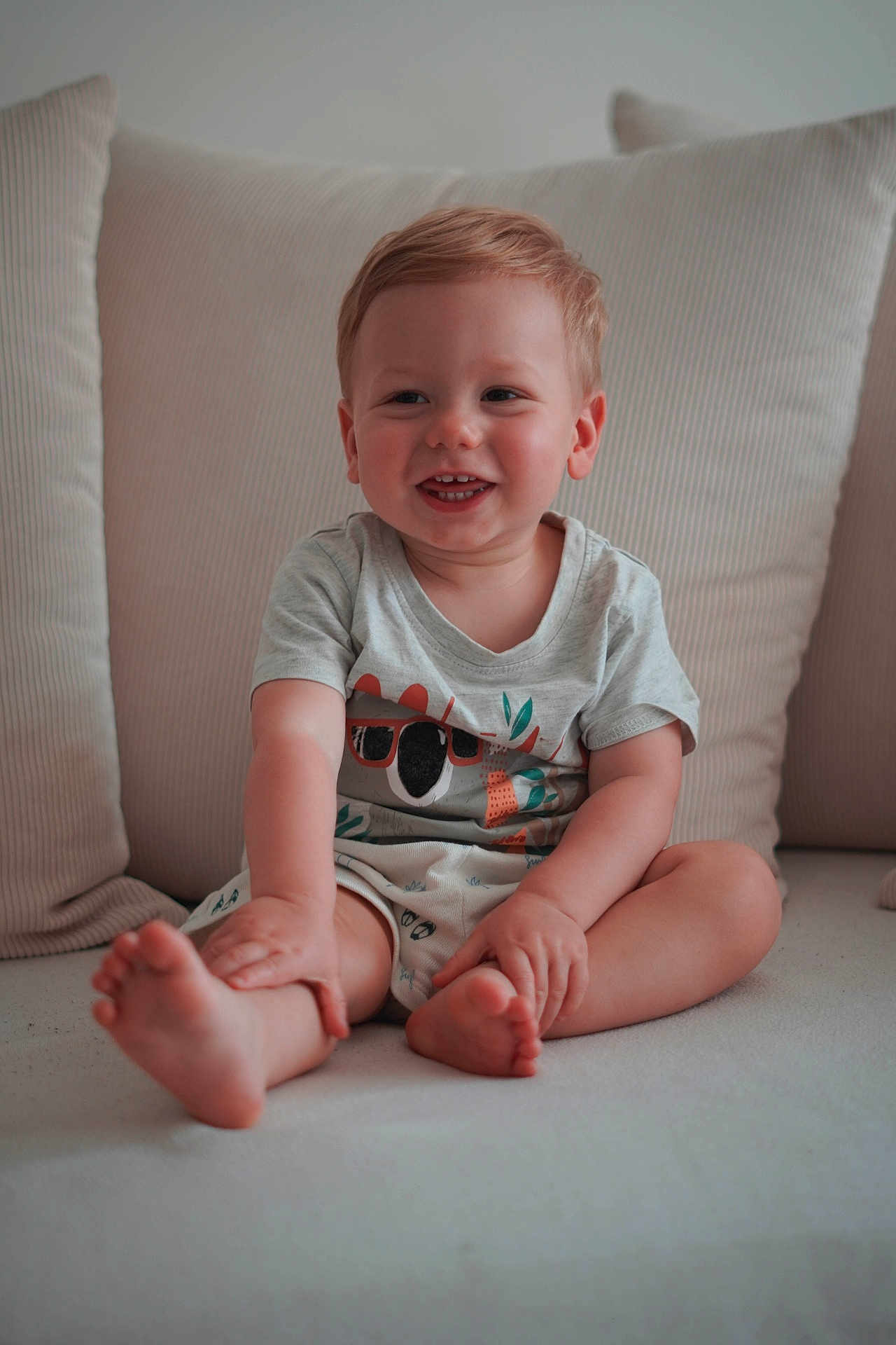 Lucas a rejoint le concours — aidez-le/la à gagner de superbes lots ! barefoot, blond_hair, child, clothing, comfort, couch, cushion, cute, face, happy, home, indoor, light_skin, portrait, shorts, sitting, smiling, toddler, tshirt, young_child