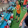 amusement_park, boy, carnival_game, cartoon_characters, casual_clothing, child, colorful_buckets, crowd, festival, fun, grass, lights, night, orange_shirt, people, play, red_balls, sandals, smile, water_trough