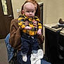 Hudson is registered to the contest to win money with this photo: baby, blanket, brown_sweater, casual, chair, child, clothing, expression, face, hands, holding, indoor, jeans, person, phone, room, sitting, socks, table, yellow_shirt