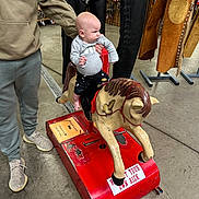 Hudson is registered to the contest to win money with this photo: adult_hand, baby, baby_clothes, child, clothing, coin_operated_ride, concrete_floor, floor, grip, indoor, leather_chaps, person, ride, shoes, stall, store, toy, toy_horse, vintage, western_gear