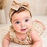 baby, infant, child, headband, bow, cute, smile, lying_down, quilt, soft_texture, indoors, portrait, closeup, skin, eyes, hand, clothing, neutral_background, adorable, happy