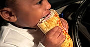 Marion is registered to the contest to win money with this photo: child, sandwich, food, eating, car, steering_wheel, white_shirt, orange_pants, hand, face, portrait, indoor, seat, dark_background, casual_clothing, young_child, appetite, candid, closeup, meal