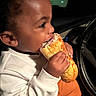 child, sandwich, food, eating, car, steering_wheel, white_shirt, orange_pants, hand, face, portrait, indoor, seat, dark_background, casual_clothing, young_child, appetite, candid, closeup, meal