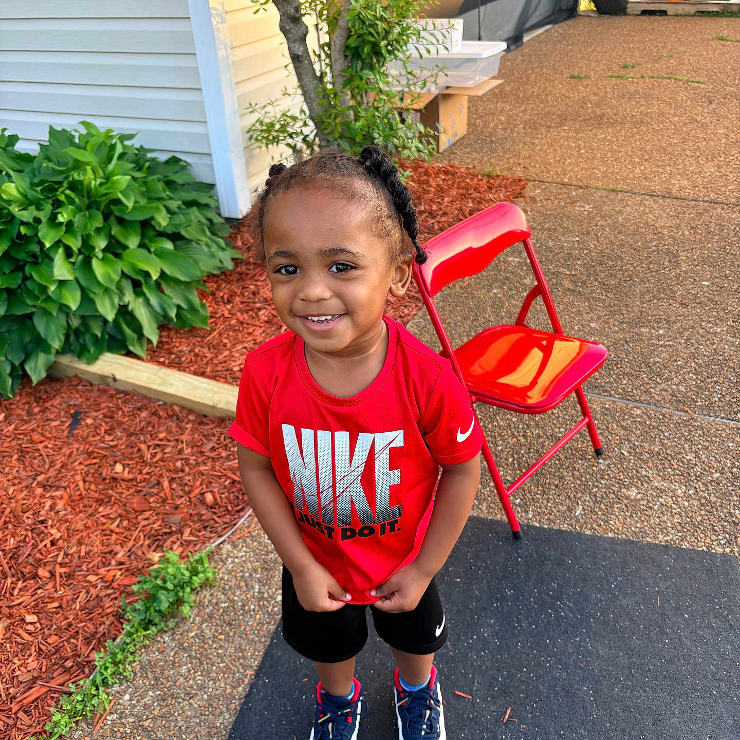 Marion is registered to the contest to win money with this photo: toddler, child, smiling, nike_shirt, red_shirt, sneakers, outdoor, red_chair, driveway, green_plants, mulch, curly_hair, sunlight, happy, person, casual_clothing, young_child, standing, daylight, garden