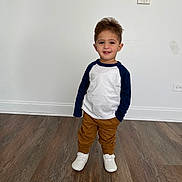 Mohammad Hussein is registered to the contest to win money with this photo: toddler, child, smiling, standing, white_shirt, navy_sleeves, brown_pants, white_shoes, wooden_floor, indoor, person, young_child, casual_clothing, happy, portrait, full_body, clean_background, wall, flooring, cute