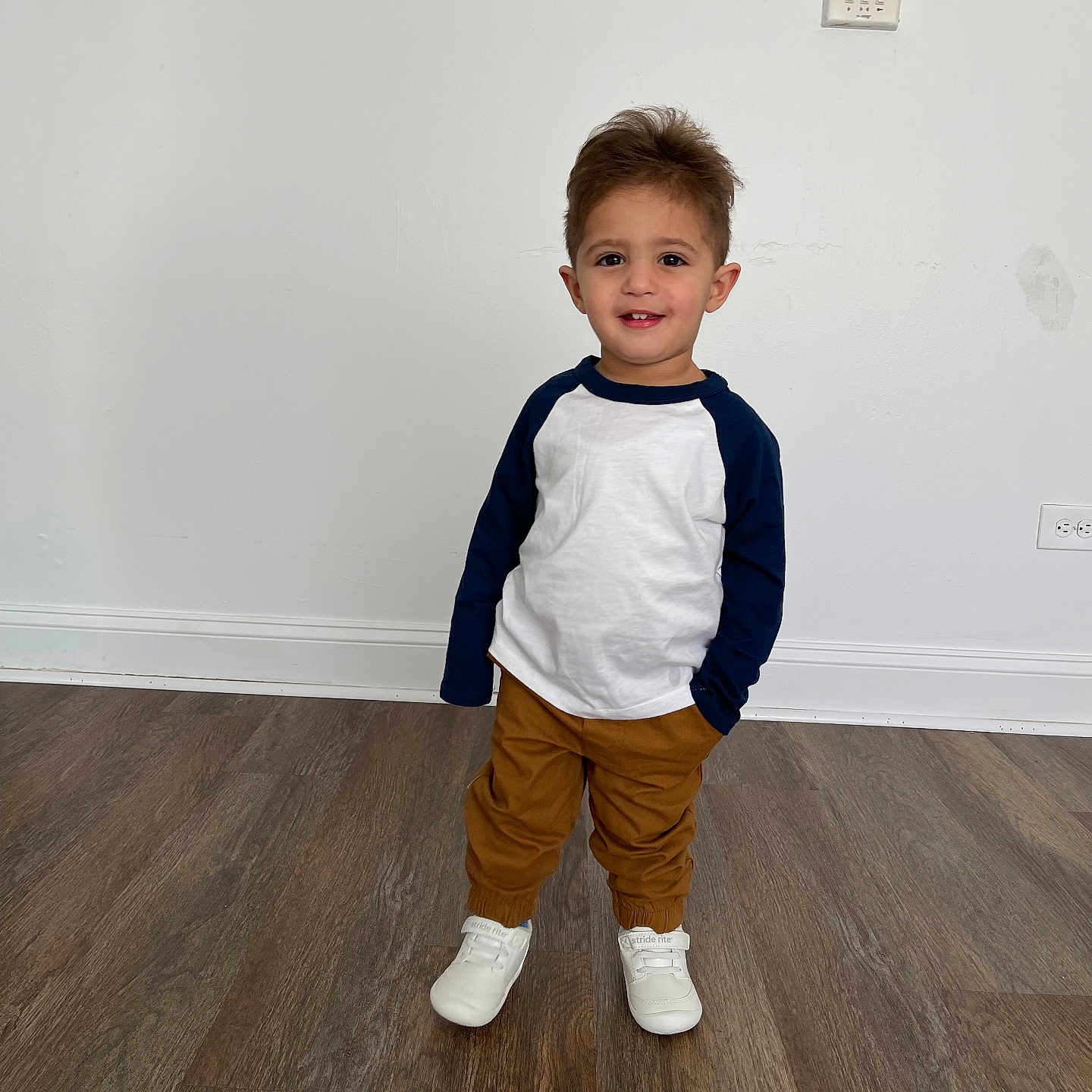 Mohammad Hussein is registered to the contest to win money with this photo: brown_pants, casual_clothing, child, clean_background, cute, flooring, full_body, happy, indoor, navy_sleeves, person, portrait, smiling, standing, toddler, wall, white_shirt, white_shoes, wooden_floor, young_child