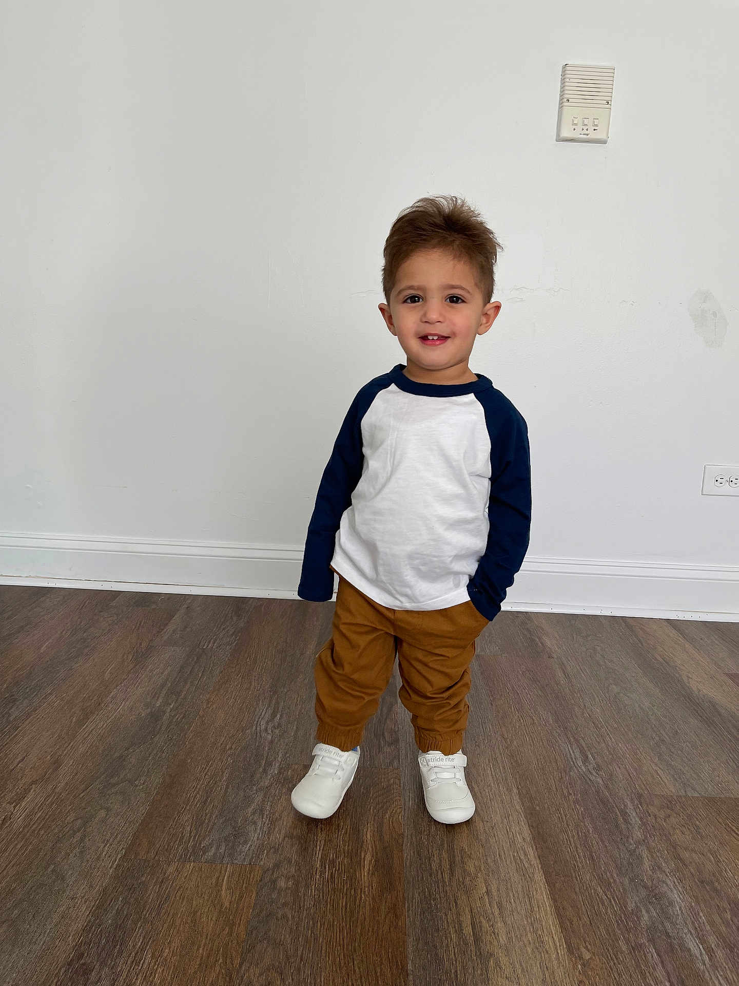 Mohammad Hussein is registered to the contest to win money with this photo: toddler, child, smiling, standing, white_shirt, navy_sleeves, brown_pants, white_shoes, wooden_floor, indoor, person, young_child, casual_clothing, happy, portrait, full_body, clean_background, wall, flooring, cute