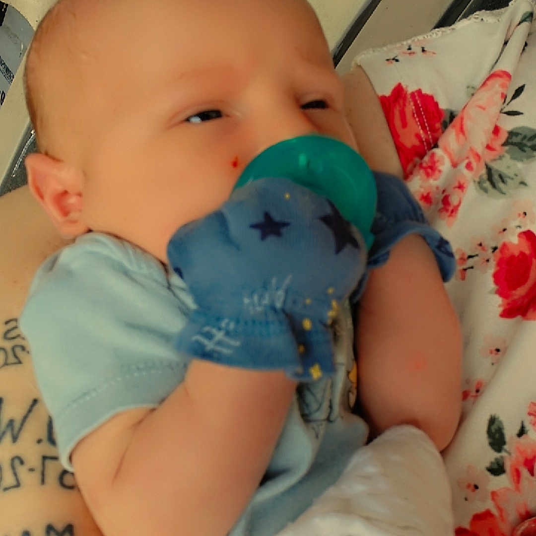 Callum is registered to the contest to win money with this photo: arm, baby, blanket, closeup, clothing, comfort, cute, fabric, floral_pattern, holding, indoor, infant, mittens, newborn, pacifier, person, resting, skin, soft, tattoo