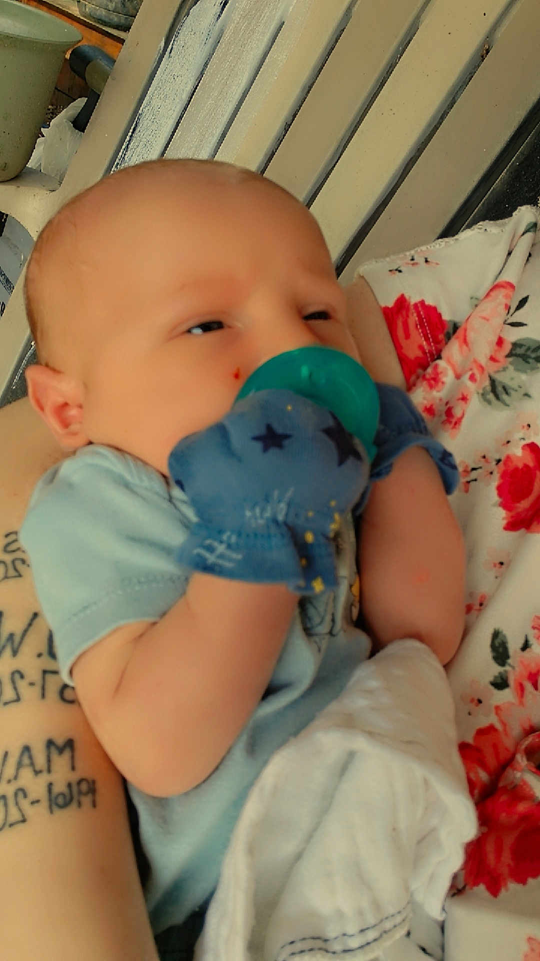 Callum is registered to the contest to win money with this photo: baby, pacifier, mittens, infant, arm, tattoo, floral_pattern, fabric, clothing, person, closeup, comfort, newborn, holding, skin, cute, resting, indoor, soft, blanket