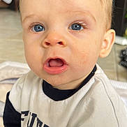 Liam a rejoint le concours — aidez-le/la à gagner de superbes lots ! toddler, child, blue_eyes, blonde_hair, superman_logo, white_shirt, navy_sleeves, indoor, curious_expression, open_mouth, portrait, young_child, cute, baby_face, casual_clothing, floor_tiles, home_interior, sitting, person, closeup