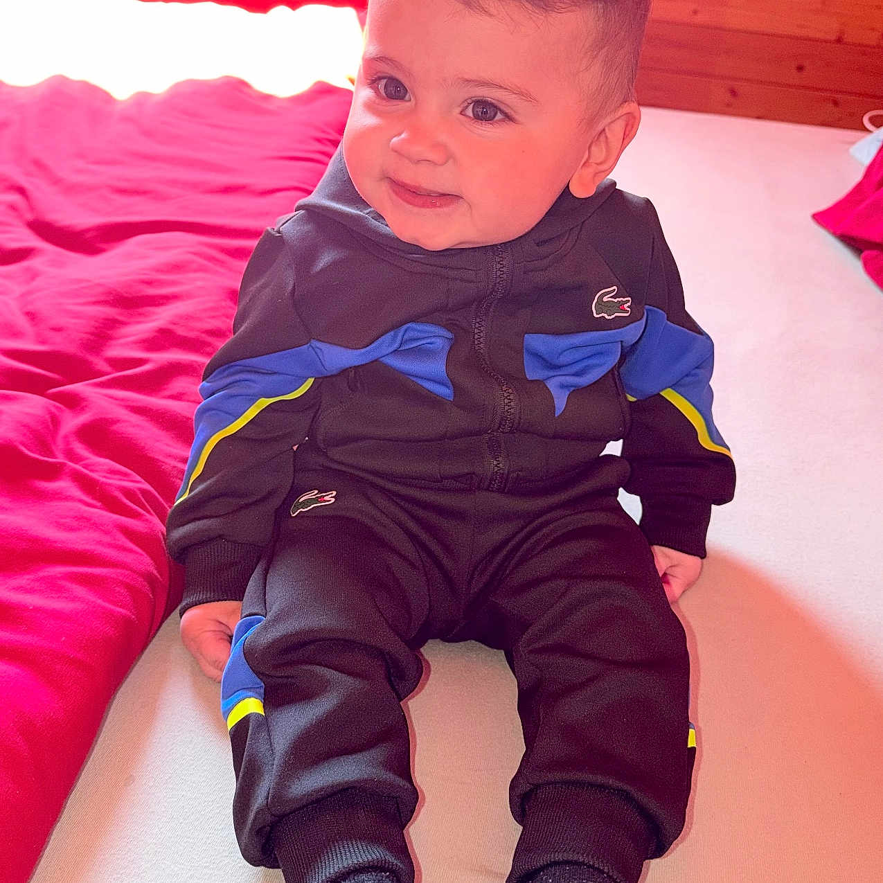Enzo participe au concours pour gagner de l'argent avec cette photo : toddler, child, bed, bedding, red_bedding, tracksuit, black_tracksuit, blue_accent, yellow_accent, socks, indoor, wooden_wall, smile, face, person, cute, seated, portrait, baby, cozy
