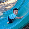 baby, child, toddler, playground, slide, outdoor, park, climbing, sitting, blue_slide, striped_shirt, black_pants, curly_hair, face, portrait, looking_at_camera, playground_equipment, wood_chips, daylight, smiling