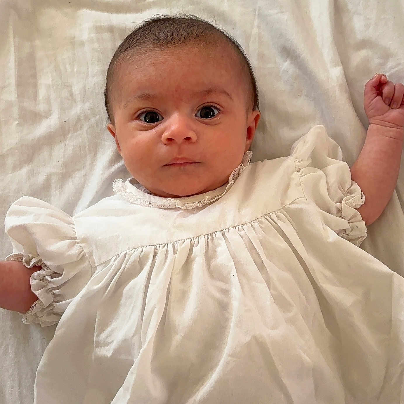Nyla is registered to the contest to win money with this photo: baby, blanket, child, curious, cute, expression, face, fists, head, indoors, infant, laying_down, newborn, portrait, skin, sleepwear, small_hands, soft_lighting, white_dress, wide_eyes