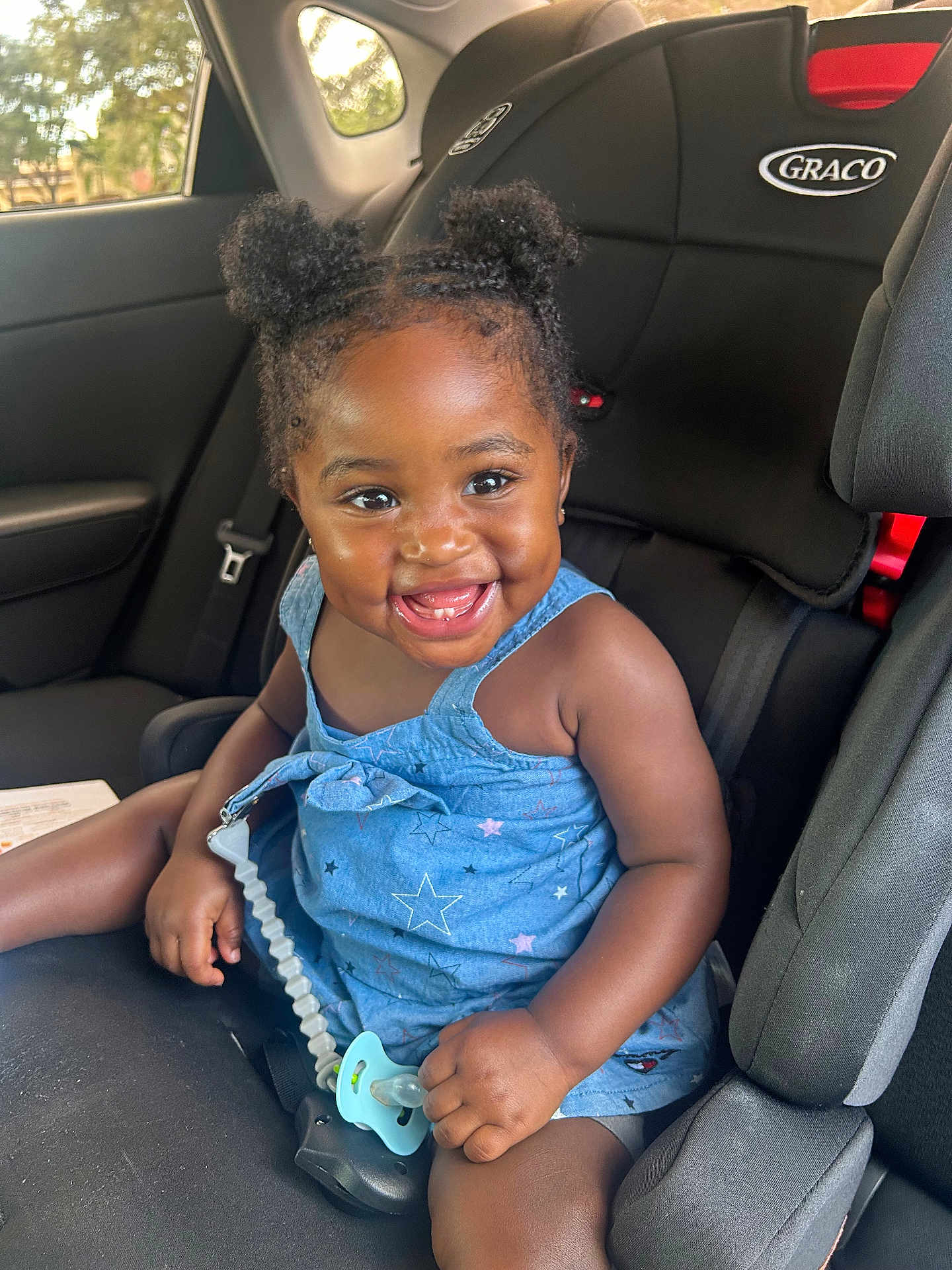 Alexis is registered to the contest to win money with this photo: toddler, child, smiling, car_seat, pacifier, blue_dress, afro_hairstyle, hair_puffs, seatbelt, car_interior, happy, cute, young_child, sitting, indoors, person, portrait, baby, joyful, seat
