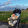 Gaspard participe au concours pour gagner de l'argent avec cette photo : baby, stroller, infant, child, grass, meadow, sky, clouds, water, shoreline, trees, path, puddle, smile, coat, winter_clothes, sunlight, outdoors, landscape, happy