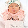 baby, newborn, infant, pink_clothing, hat, blanket, embroidered_text, face, hands, portrait, cute, child, soft_texture, sleepwear, indoor, lying_down, expression, closeup, skin, headwear