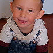 Naël participe au concours pour gagner de l'argent avec cette photo : child, boy, smile, cap, baseball_cap, denim_overalls, hoodie, happy, indoors, floor, tile_floor, casual_clothing, portrait, sitting, cute, young, face, headwear, person, playful