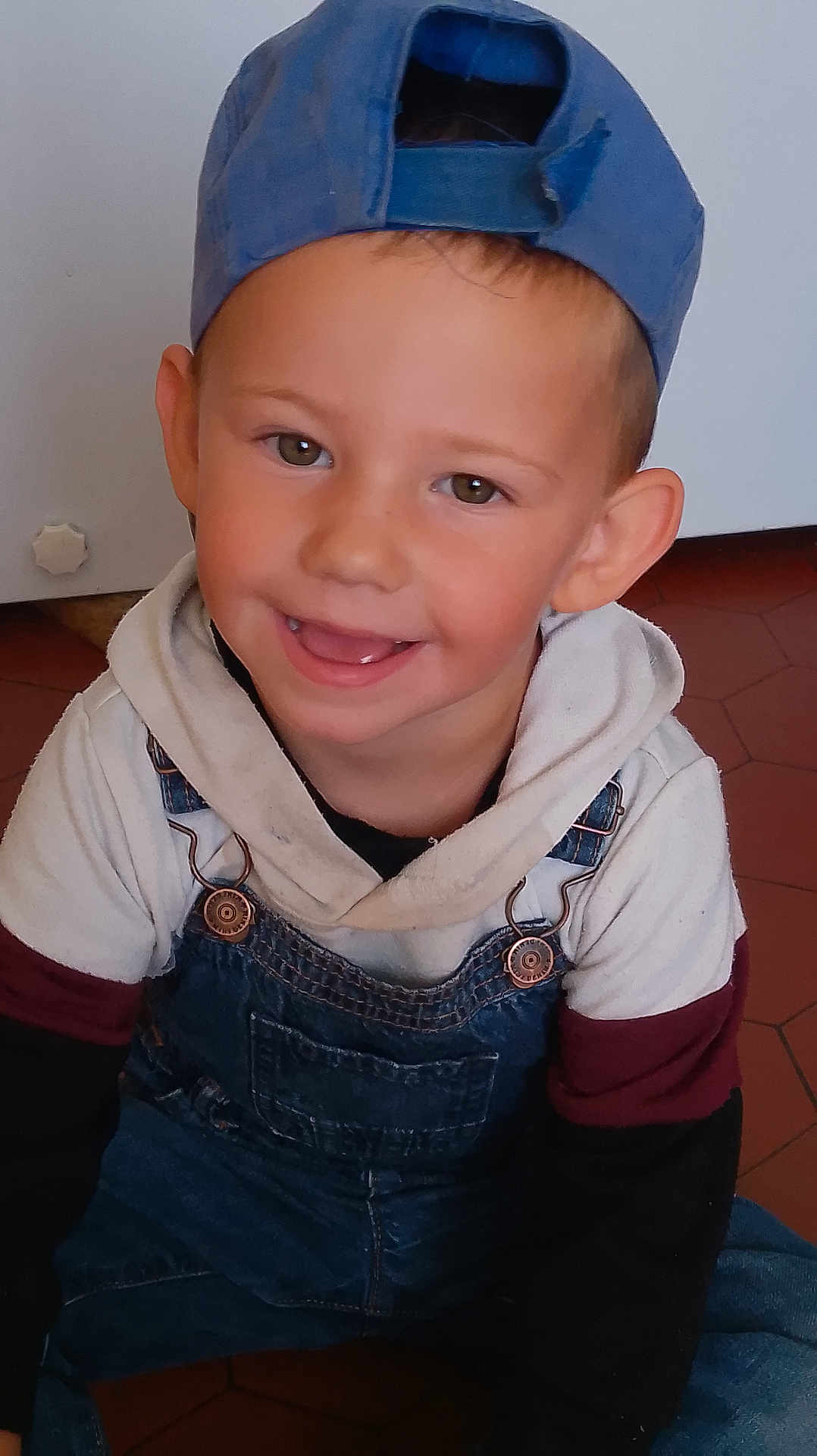 Naël participe au concours pour gagner de l'argent avec cette photo : child, boy, smile, cap, baseball_cap, denim_overalls, hoodie, happy, indoors, floor, tile_floor, casual_clothing, portrait, sitting, cute, young, face, headwear, person, playful