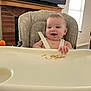 baby, high_chair, smiling, crumbs, indoor, living_room, television, window, wooden_mantel, seatbelt, child, person, furniture, happy, face, hand, clothing, infant, home, daylight