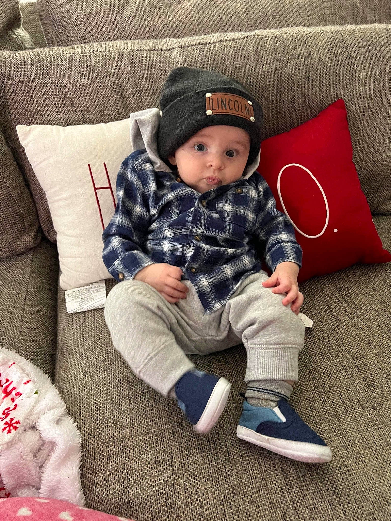 Lincoln joined the competition — help win amazing prizes! baby, baby_toddler_clothing, cap, child, comfort, couch, face, hat, headwear, human_leg, lap, linens, living_room, pattern, person, sitting, sleeve, sock, surprise, throw_pillow