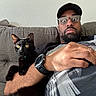 Kenobi joined the competition — help win amazing prizes! man, cat, black_cat, glasses, cap, couch, arm, watch, facial_hair, indoor, furniture, wide_eyes, surprised, pet, relaxing, casual, person, animal, companion, home