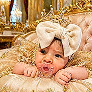 Laura joined the competition — help win amazing prizes! baby, child, pacifier, crown, bow, golden, luxury, plush, cushion, royalty, dress, sparkle, chandelier, indoor, portrait, cute, infant, decor, face, hands