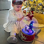 Lyana participe au concours pour gagner de l'argent avec cette photo : celebration, child, christmas_tree, curious, dress, festive, floor, happy, holiday_decor, indoors, lights, ornaments, reindeer_headband, seasonal, shoes, sitting, snow_globe, toddler, toy, winter