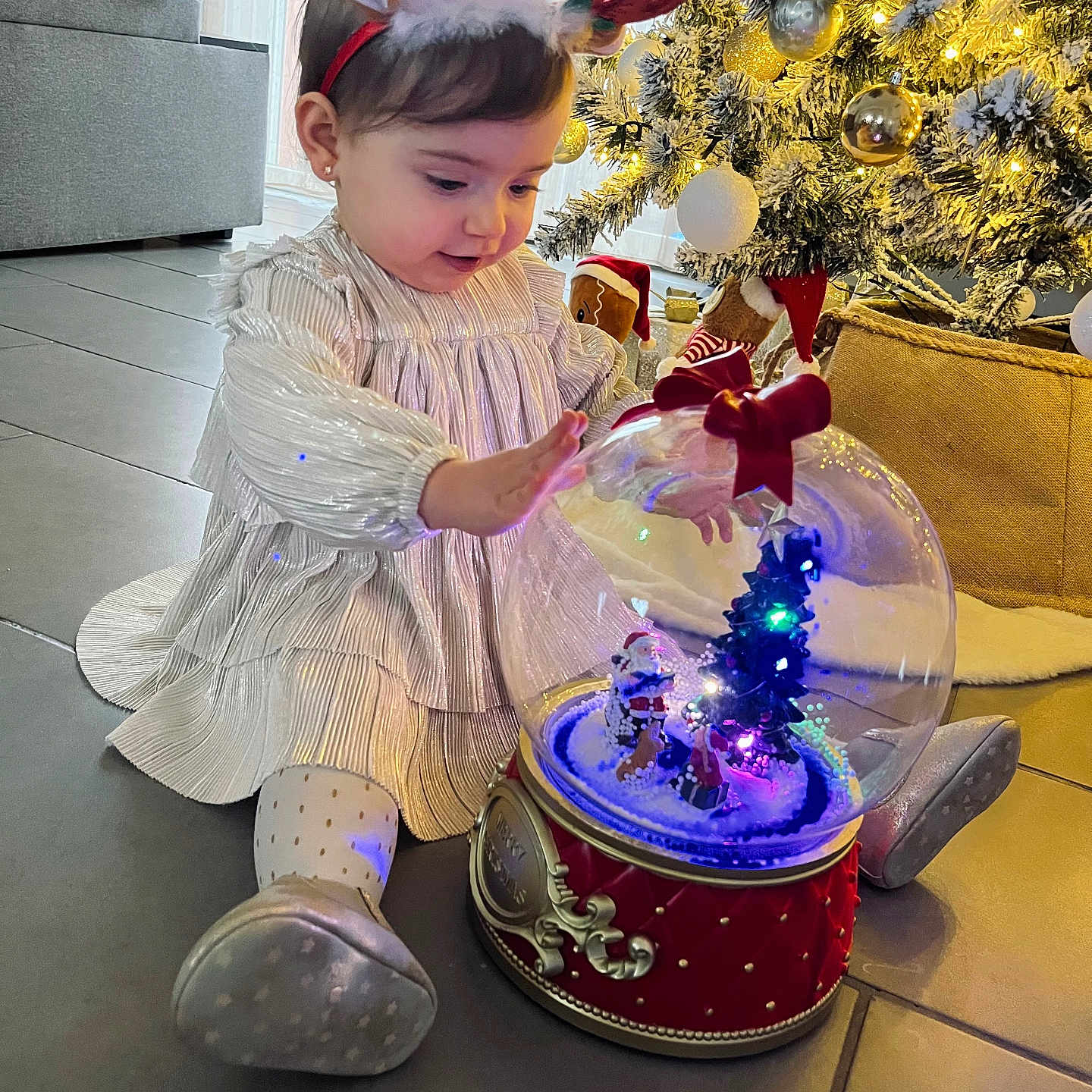 Lyana participe au concours pour gagner de l'argent avec cette photo : celebration, child, christmas_tree, curious, dress, festive, floor, happy, holiday_decor, indoors, lights, ornaments, reindeer_headband, seasonal, shoes, sitting, snow_globe, toddler, toy, winter