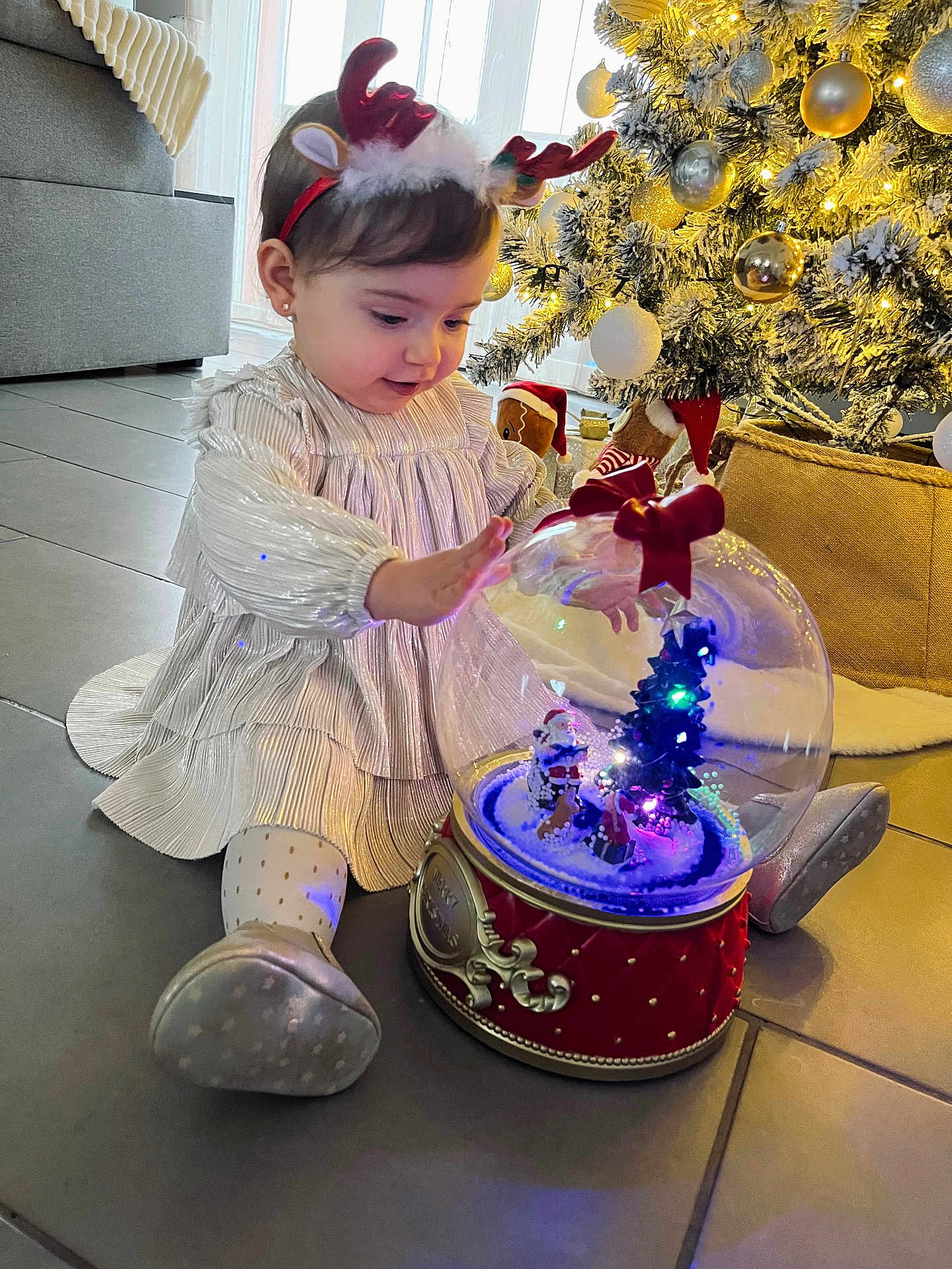 Lyana participe au concours pour gagner de l'argent avec cette photo : celebration, child, christmas_tree, curious, dress, festive, floor, happy, holiday_decor, indoors, lights, ornaments, reindeer_headband, seasonal, shoes, sitting, snow_globe, toddler, toy, winter