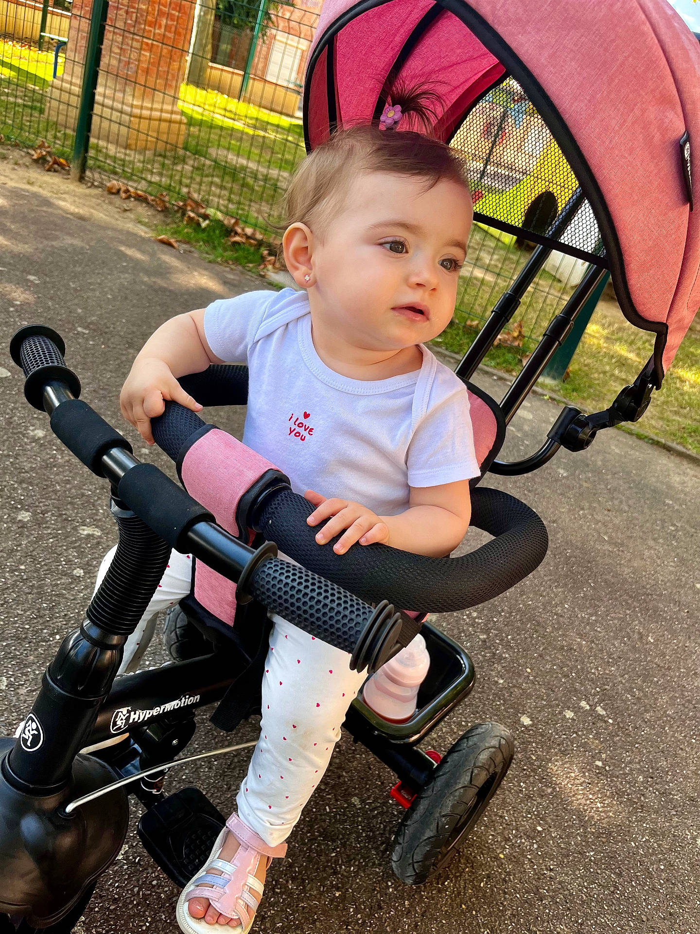 Lyana participe au concours pour gagner de l'argent avec cette photo : baby, child, curious, cute, fence, grass, outdoor, park, pavement, person, pink, playground, sandals, sitting, small_pony_tail, sunlight, toddler, toy, tricycle, white_clothing