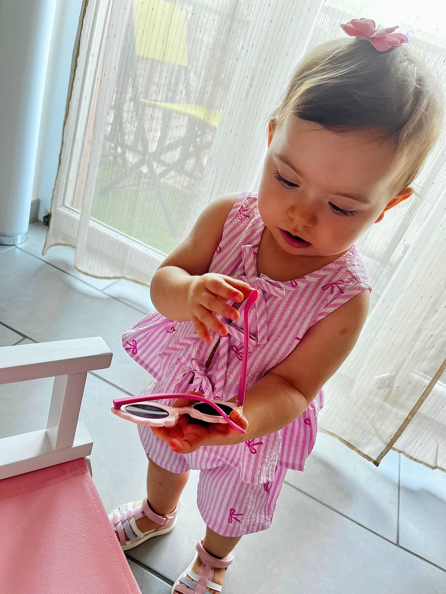 Lyana participe au concours pour gagner de l'argent avec cette photo : child, curious, curtains, hair_accessory, home, indoor, looking_down, person, pink_clothing, pink_cushion, shoes, standing, striped_outfit, summer_outfit, sunglasses, sunlight, tile_floor, toddler, white_chair, window