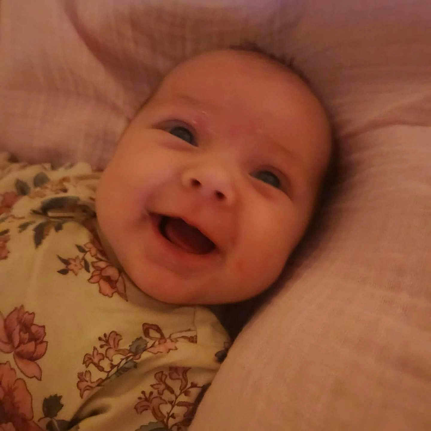Sylvia is registered to the contest to win money with this photo: baby, smiling, face, infant, cushion, pink, floral, clothing, happy, portrait, child, young, cute, head, closeup, lying, soft, comfort, indoors, person