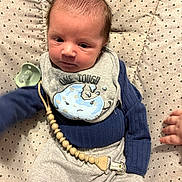 Bailor is registered to the contest to win money with this photo: baby, newborn, infant, bib, pacifier, pacifier_clip, wooden_beads, blanket, lounger, patterned_fabric, blue_shirt, grey_pants, hand, face, head, hair, portrait, indoor, cute, sleepy